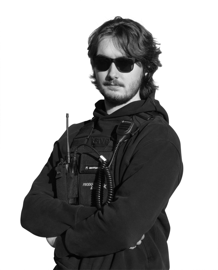 Man wearing a black hoodie and tactical vest with a radio, arms crossed and looking at the camera, wearing sunglasses.