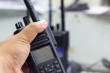 A close-up shot of a person's hand firmly gripping a modern black two-way radio with an antenna and screen, ready for communication. Blurry background shows more communication devices
